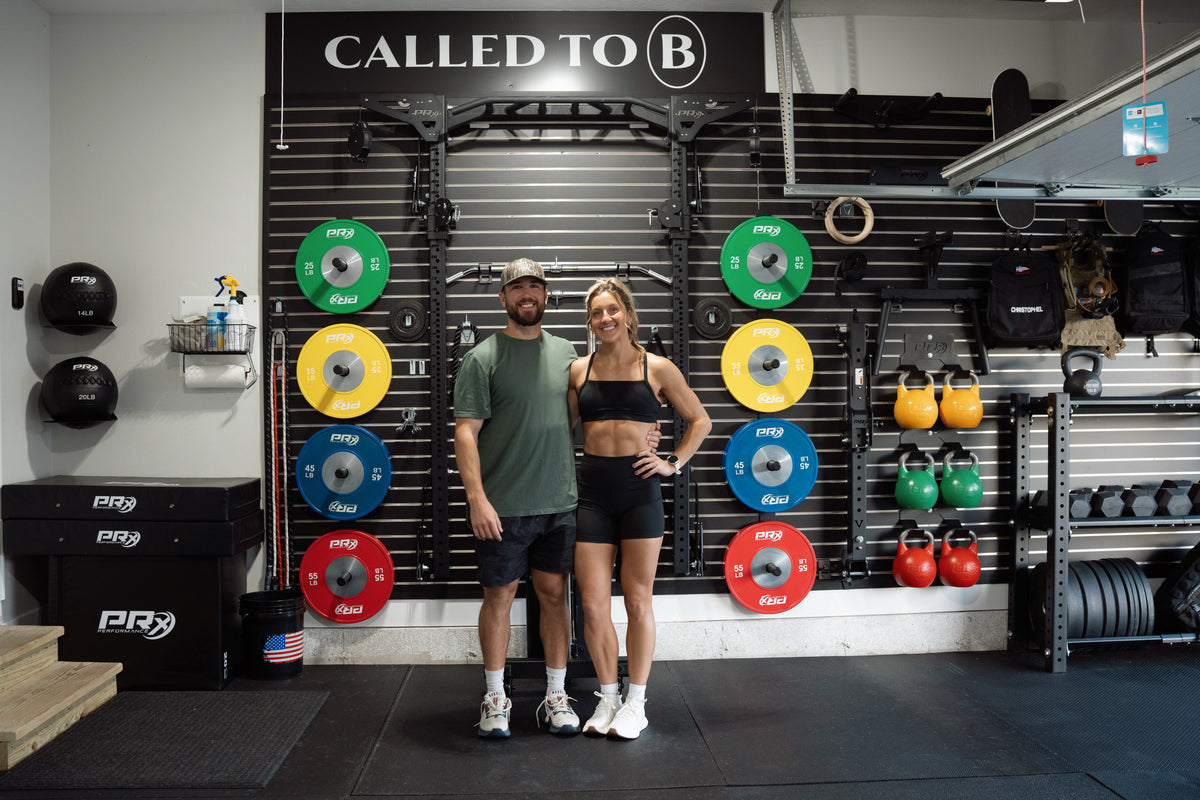 A man and a woman stand in a gym in front of a branded wall with barbell plates and kettlebells. The man wears a green t-shirt and shorts, and the woman wears a black sports bra and shorts.