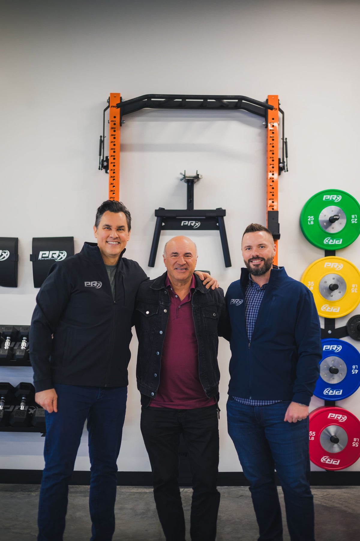 Three men standing together in a gym setting with fitness equipment in the background.