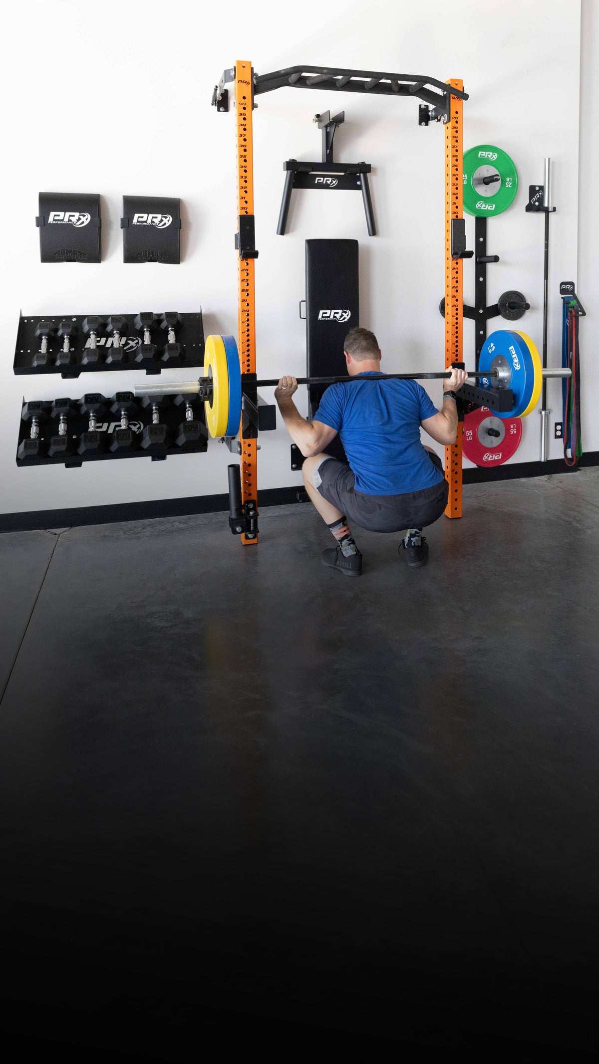 A man in a blue shirt and gray shorts squats inside a black and orange power rack loaded with colorful weight plates in a gym setting.