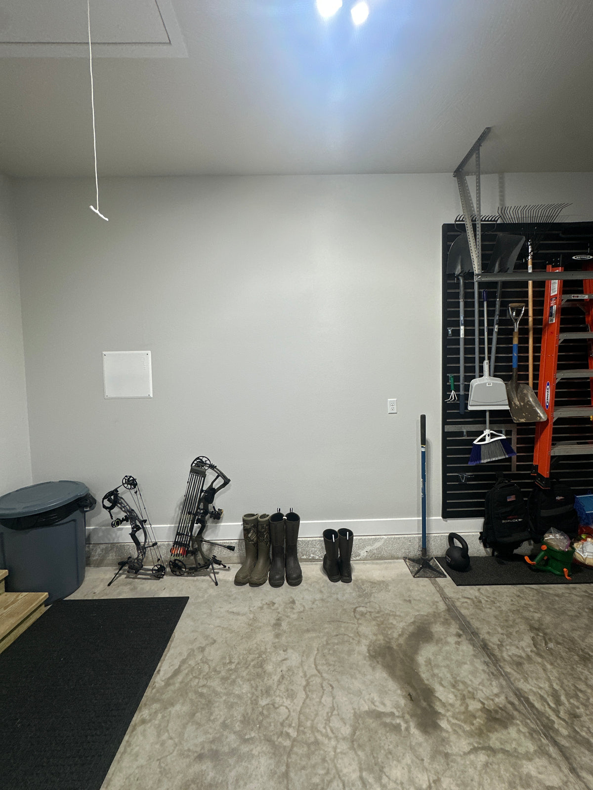 A garage interior with a gray wall and concrete floor, showing a set of bowfishing rods leaning against the wall, a pair of boots, and a black shelving unit holding various fishing and storage items.