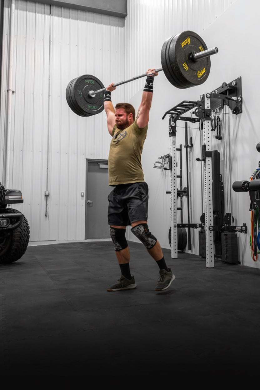 A man in a gym setting, wearing a green t-shirt, black shorts, and knee sleeves, lifting a barbell overhead. The background shows a white wall with various gym equipment, including a pull-up bar, weight plates, and a resistance machine, as well as a black rubber floor mat.