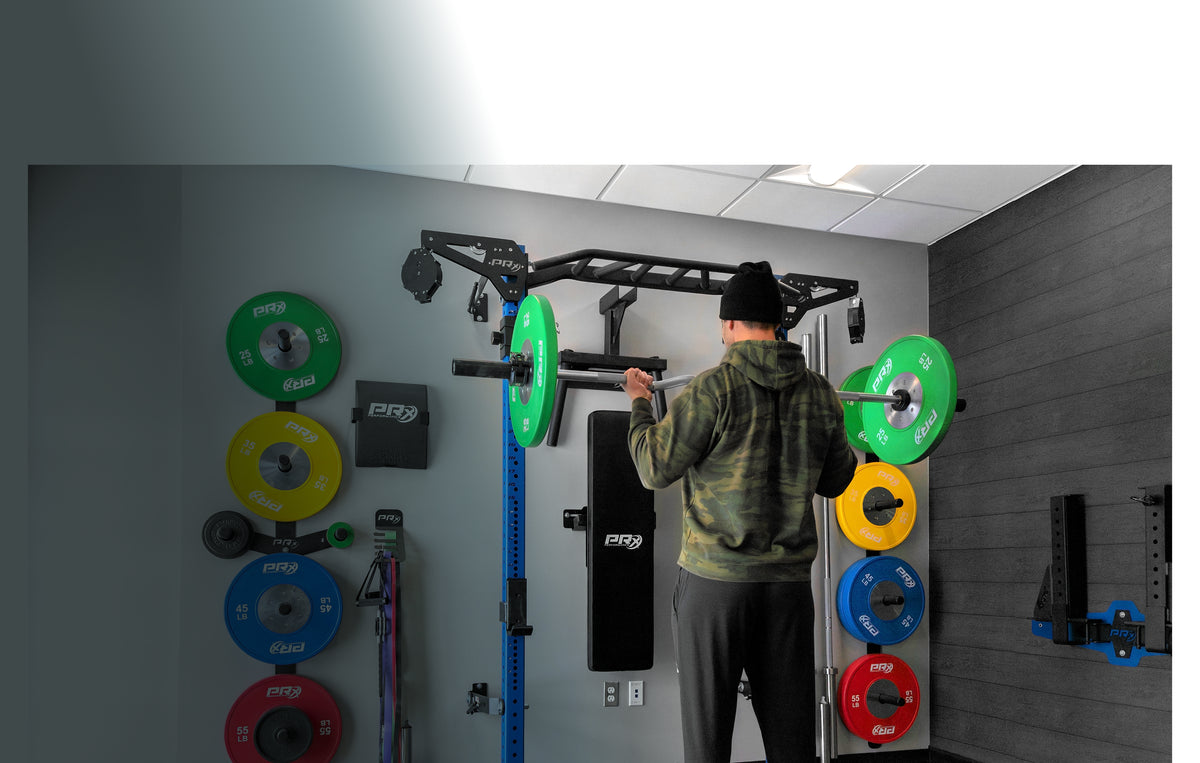 A man in a camouflage hoodie and black beanie stands facing a black wall-mounted pull-up bar and weight rack system in a home gym. Colorful weight plates are stacked on the left and right walls, and the man is shown from behind.
