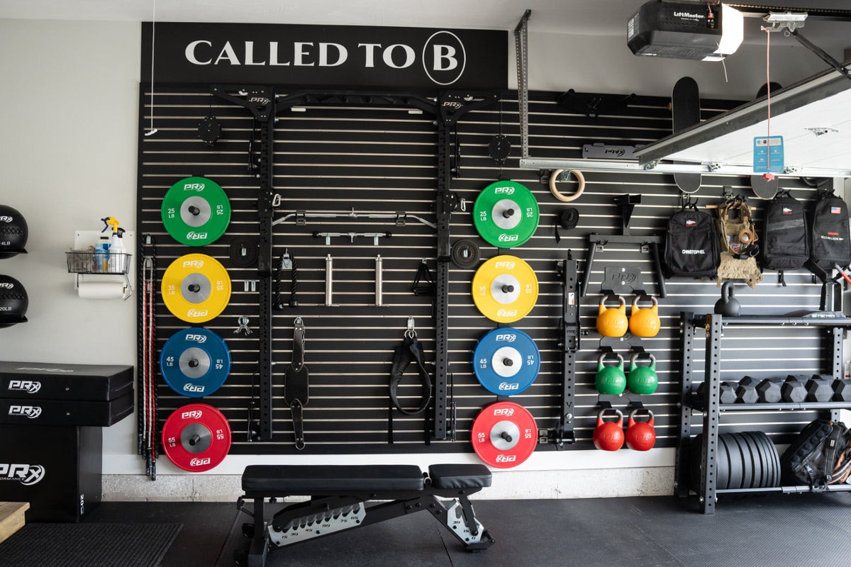A home gym interior with a black slat-wall fitness system holding colorful weight plates, pull-up bars, straps, hanging bags, a bench, and racks of dumbbells. A large sign above reads 'CALLED TO B'.