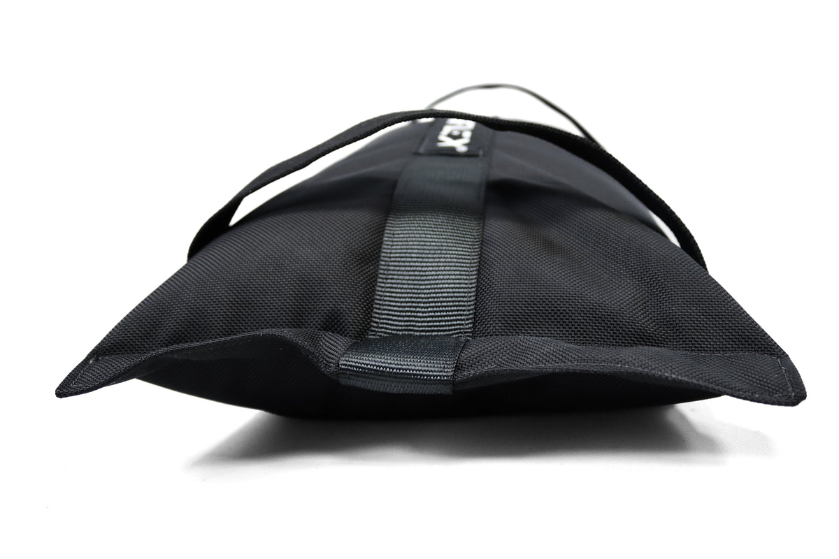 A black textured canvas sandbag with a black webbing strap and a small white logo patch, shown on a clean white background.
