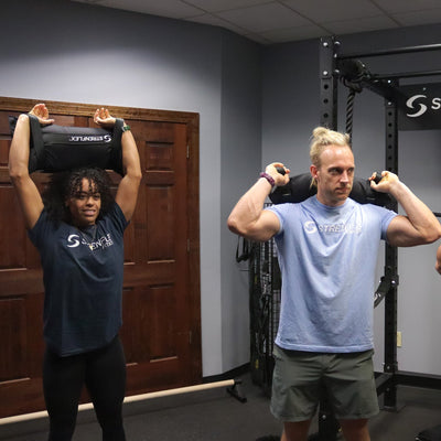 Three people in a gym setting performing squats while carrying black Sandbagz weight bags on their shoulders. A Strenflex Fitness power rack is visible in the background.