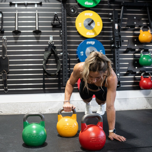 A woman in a black tank top and white leggings does a push-up in a gym while holding a yellow kettlebell. Three kettlebells in green, yellow, and red are on the floor in front of her, with weight plates and gym equipment hanging on a black slatwall behind her.